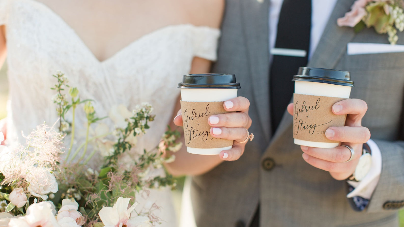 Bride and groom holding coffee cups with custom wedding sleeves, surrounded by floral arrangements.