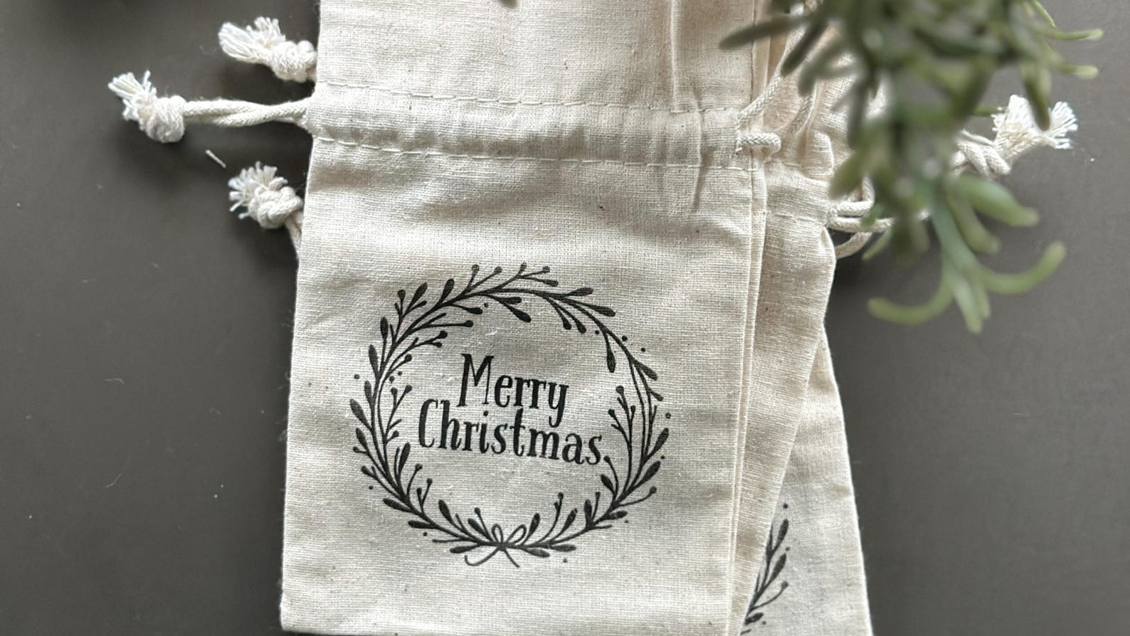 Natural cotton drawstring bags with a Merry Christmas wreath graphic printed on them surrounded by greenery.
